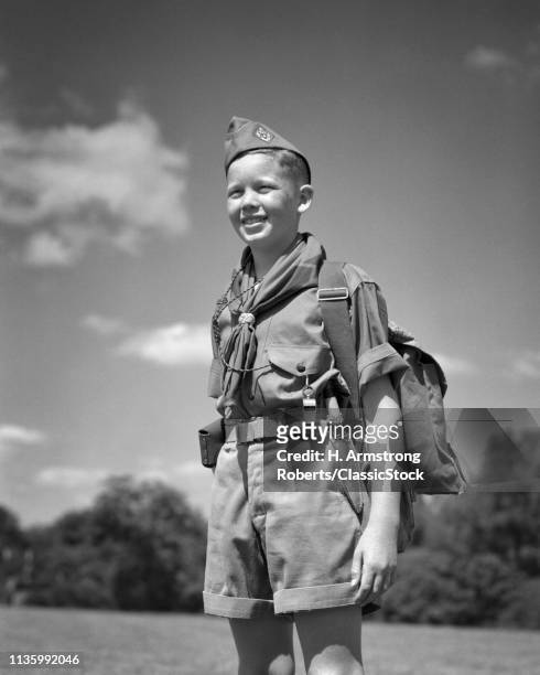 1950s SMILING YOUNG TEENAGE BOY SCOUT STANDING AT ATTENTION WEARING UNIFORM CAP SHIRT NECKERCHIEF SHORTS PACK WHISTLE OUTDOORS