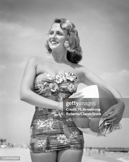 1950s PORTRAIT OF SMILING BLOND WOMAN IN PRINT ONE PIECE BATHING SUIT STANDING ON BEACH AT SEASHORE HOLDING PLASTIC BEACH BALL