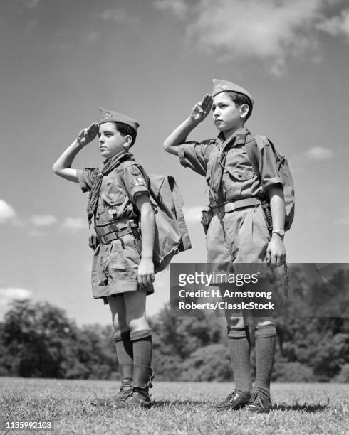 1950s TWO BOY SCOUTS WEARING SUMMER UNIFORMS STANDING AT ATTENTION EQUIPPED WITH BACKPACKS THREE FINGER HAND SALUTING