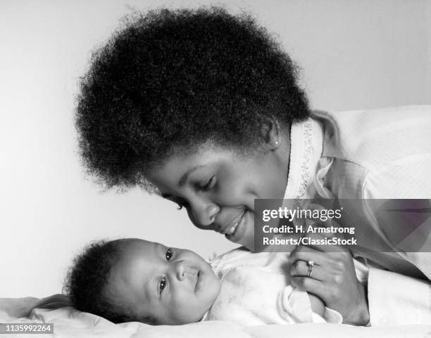 1970s SMILING AFRICAN AMERICAN MOTHER HOLDING HAND AND LOOKING AT HAPPY BABY DAUGHTER LYING ON HER BACK