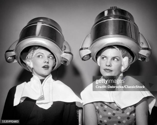 1950s TWO WOMEN SITTING UNDER BEAUTY SALON HAIR DRYERS WEARING HAIRNETS TOWELS TALKING GOSSIP