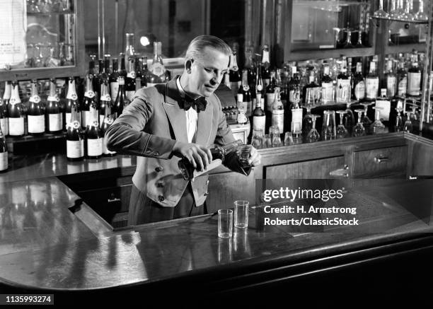 1940s 1950s MAN BARTENDER BEHIND BAR POURING A SHOT MIXING DRINK WEARING BOW TIE UNIFORM SHORT JACKET SET UP BOTTLES ON BACKBAR