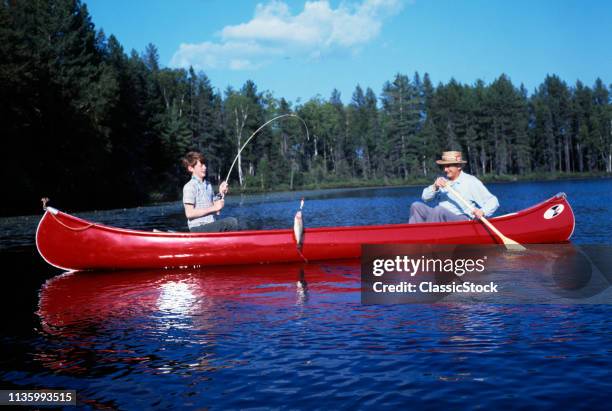 1960s FATHER AND SON FISHING TOGETHER IN RED CANOE ON RURAL LAKE BOY REELING IN FISH