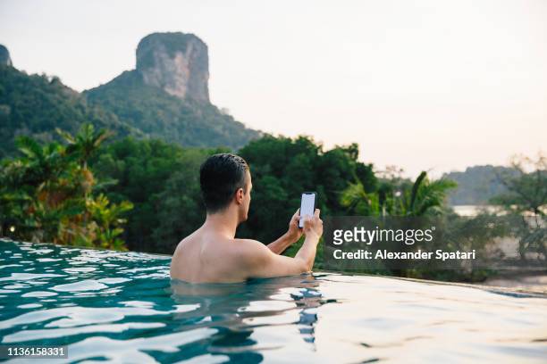 young man using smartphone in infinity pool overlooking tropical landscape - business man looking at smart phone stock-fotos und bilder