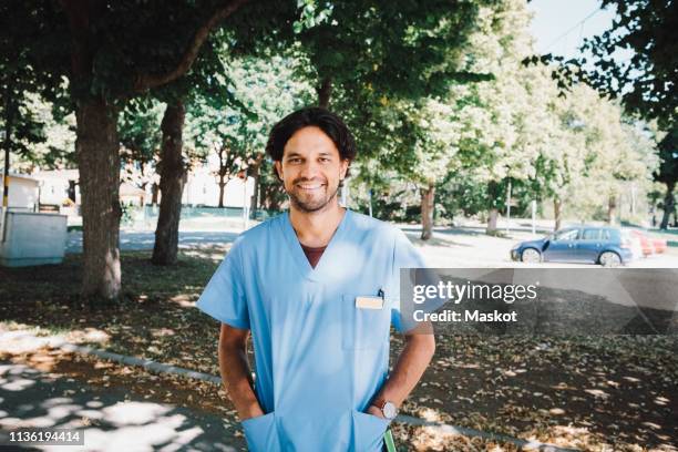 portrait of smiling male nurse standing with hands in pockets at nursing home - pflegedienst blau stock-fotos und bilder