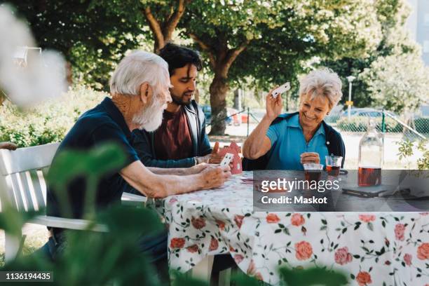 cheerful senior woman playing cards with males at table in back yard - floral pattern suit stock pictures, royalty-free photos & images