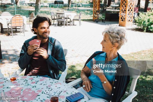 retired senior woman playing cards with smiling male caretaker sitting at table in back yard - volunteer aged care stock pictures, royalty-free photos & images