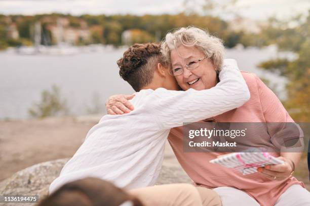 grandson embracing smiling grandmother while receiving gift on lakeshore in park during picnic - boy gift stock-fotos und bilder