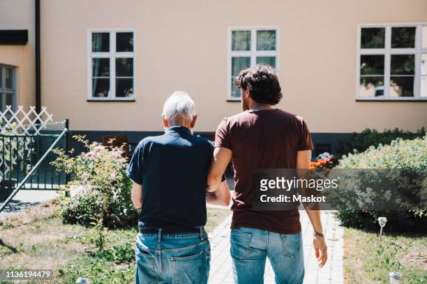 rear view of male caretaker walking arm in arm with senior man at nursing home - bidello foto e immagini stock