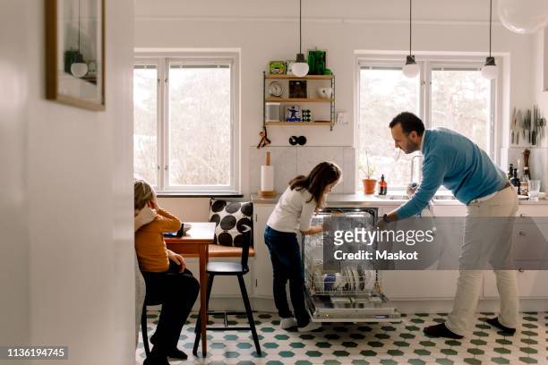 father and daughter arranging utensils in dishwasher while standing at kitchen - routine foto e immagini stock