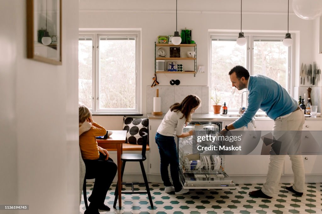Father and daughter arranging utensils in dishwasher while standing at kitchen