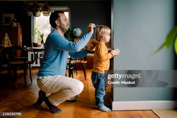 full length of father measuring daughter's height against wall at home - kinderheim stock-fotos und bilder