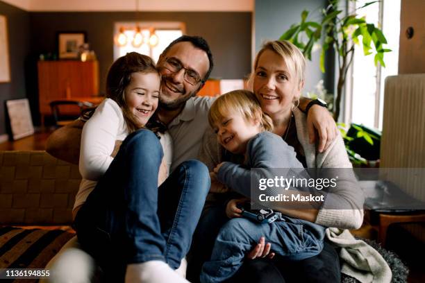 portrait of parents with cheerful daughters sitting in living room at home - sibling stock photos et images de collection