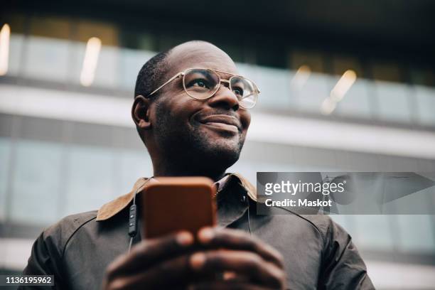 low angle view of smiling businessman using smart phone looking away while standing against building in city - modern maturity center foto e immagini stock