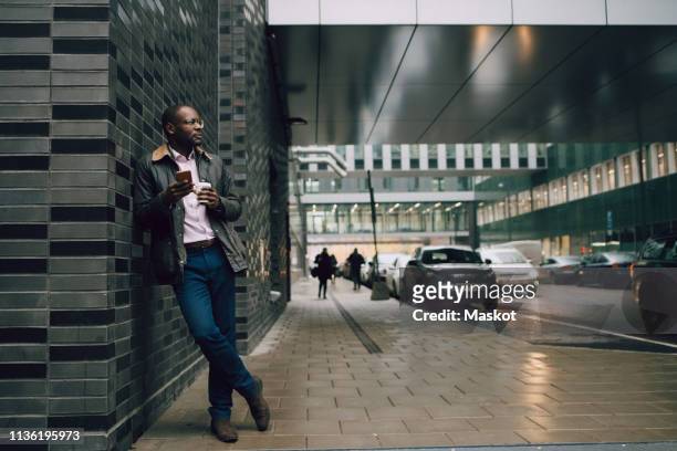 full length of businessman holding coffee and mobile phone looking away while standing on road in city - guy with phone full image ストックフォトと画像