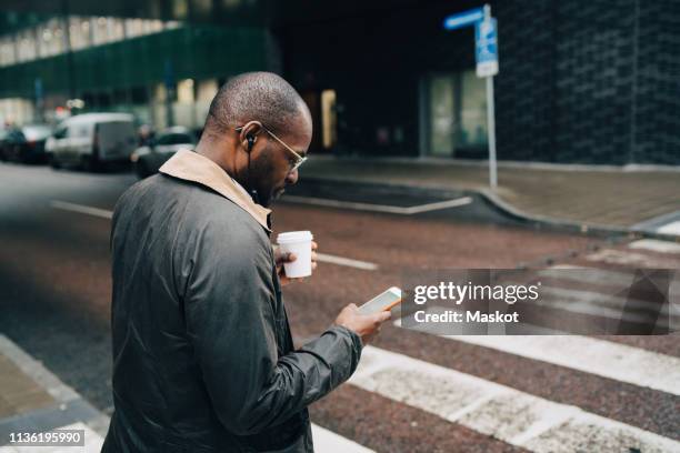 side view of businessman with coffee using mobile phone while crossing road in city - paso de cebra fotografías e imágenes de stock