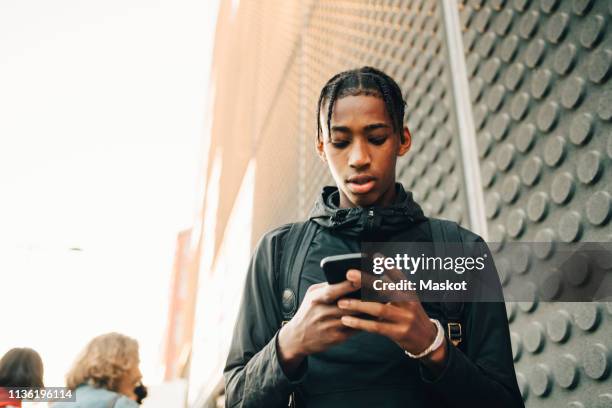 low angle view of teenage boy using mobile phone while standing in city - 3 teenagers mobile outdoors stockfoto's en -beelden