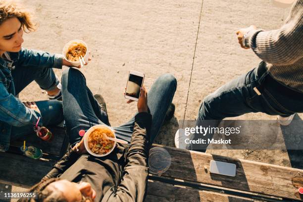 high angle view of teenage boy using smart phone while eating meal with friends on street in city - 3 teenagers mobile outdoors stock-fotos und bilder