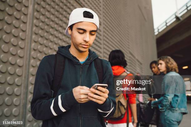 young man using mobile phone with friends standing in background on footpath - arab teen photos et images de collection