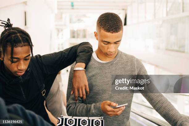 teenage boy using mobile phone while standing by friend in shopping mall - teenager boy shopping bildbanksfoton och bilder