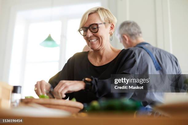 smiling mature woman preparing food with friend in background at home - mature adult cooking stock pictures, royalty-free photos & images