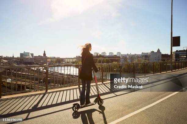 Full length of female commuter riding electric push scooter on bridge in city against sky