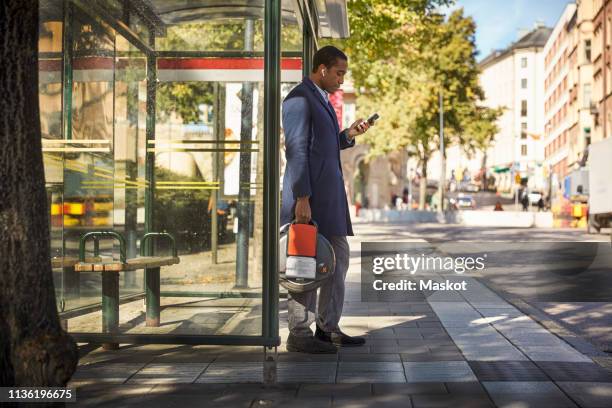 full length side view of young male commuter using smart phone while holding electric unicycle at bus stop in city - bus shelter ストックフォトと画像