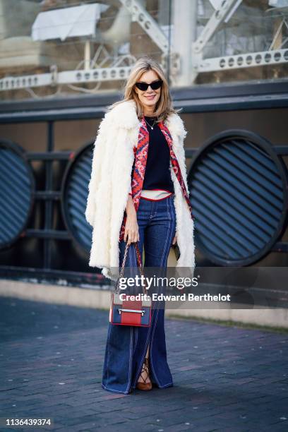 Guest wears sunglasses, a white fluffy coat, a navy blue top, a red and blue scarf, blue front split flare denim pants, tan color shoes, a white, red...