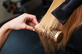 Female client in beauty salon. Close-up of hairdressers hand drying blond hair with hair dryer and round brush, doing new hairstyle
