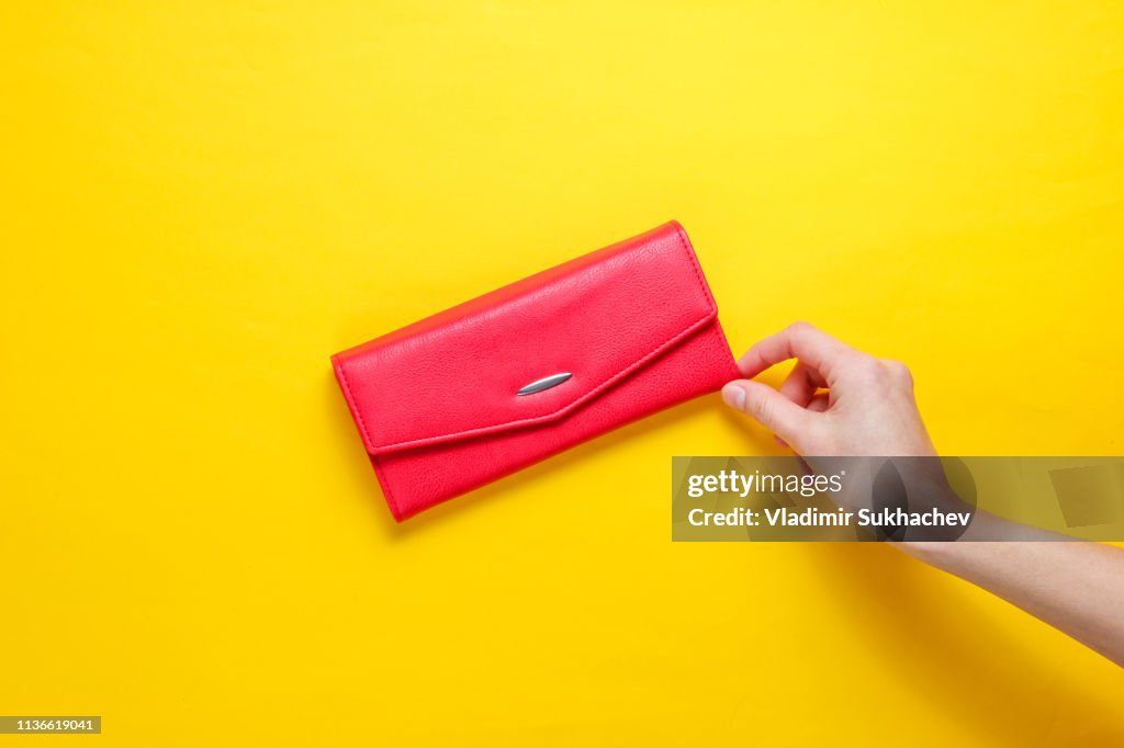 Tendência do minimalismo. A mão fêmea toma a carteira de couro vermelha no fundo amarelo. Vista superior