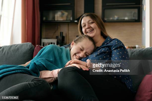 Cheerful excited friendly plus-size mother without makeup and her daughter sitting on sofa at home and laughing at movie while watching TV