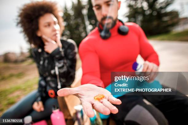 junger athletischer mann hält ein paar kapseln auf der handfläche, während er auf einer parkbank neben einem weibchen sitzt - exercise pill stock-fotos und bilder