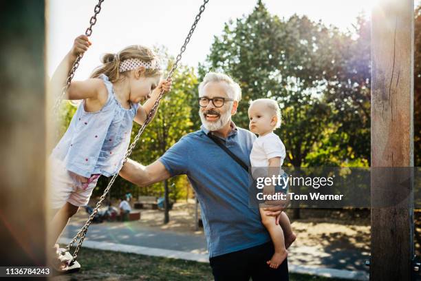 grandfather having fun with grandchildren on swings - public park playground stock pictures, royalty-free photos & images