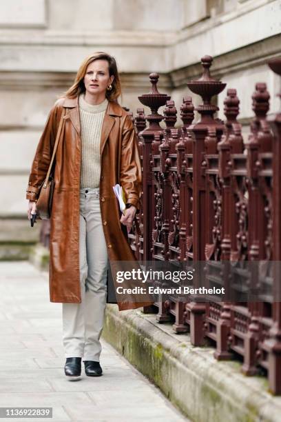 Guest wears earrings, a beige sweater, a tan color leather coat, a tan color bag, beige pants, black pointy boots, during London Fashion Week...