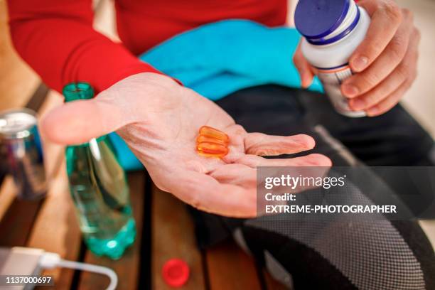 junger athletischer mann hält ein paar orange farbige kapseln auf der handfläche, während er auf einer parkbank sitzt - exercise pill stock-fotos und bilder