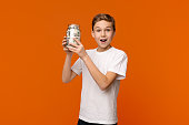 Emotional boy holding glass money box on orange background
