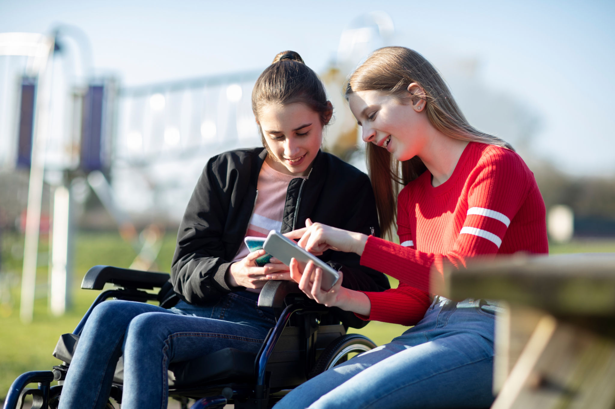 Teenage Girl In Wheelchair Looking At Mobile Phone With Friend In Park Teenage Girl In Wheelchair Looking At Mobile Phone With Friend In Park