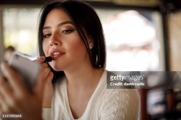 young woman applying lipstick at a cafe - woman applying makeup stock pictures, royalty-free photos & images