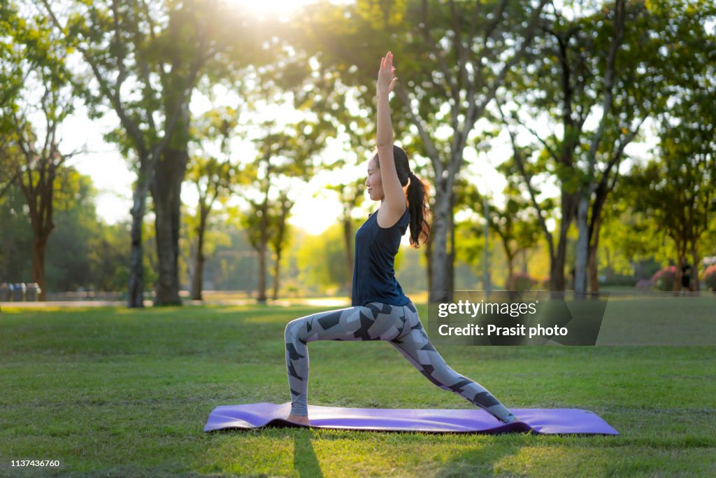 Young Asian woman practicing warrior 1 yoga in park at sunset.