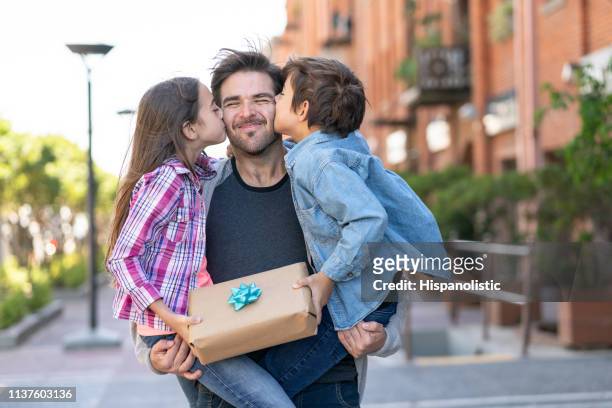 amorevole figlio e figlia che baciano papà sulla guancia mentre li porta entrambi con un regalo per la festa del papà - festa del papà foto e immagini stock