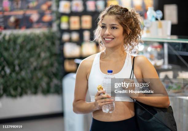 jovencita en el gimnasio comiendo un bar de cereales mientras miraba muy pensativo pero sonriendo - cereal bar fotografías e imágenes de stock