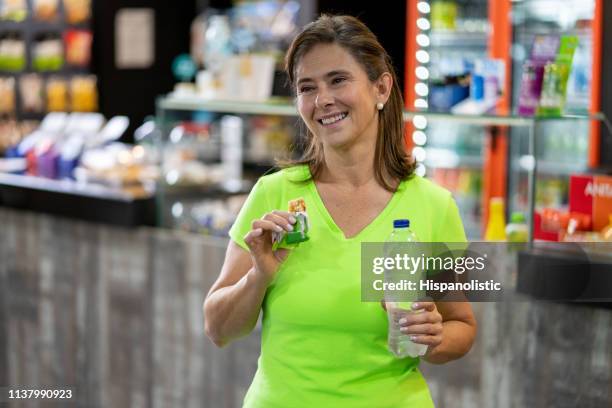 hermosa mujer madura en el gimnasio comiendo un bar de cereales y beber agua después de hacer el trabajo - cereal bar fotografías e imágenes de stock