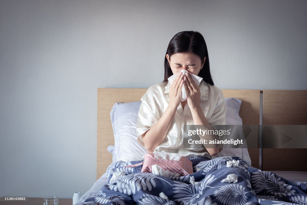 Asian woman have a cold, sitting on cozy bed using tissue for snot. sick at home