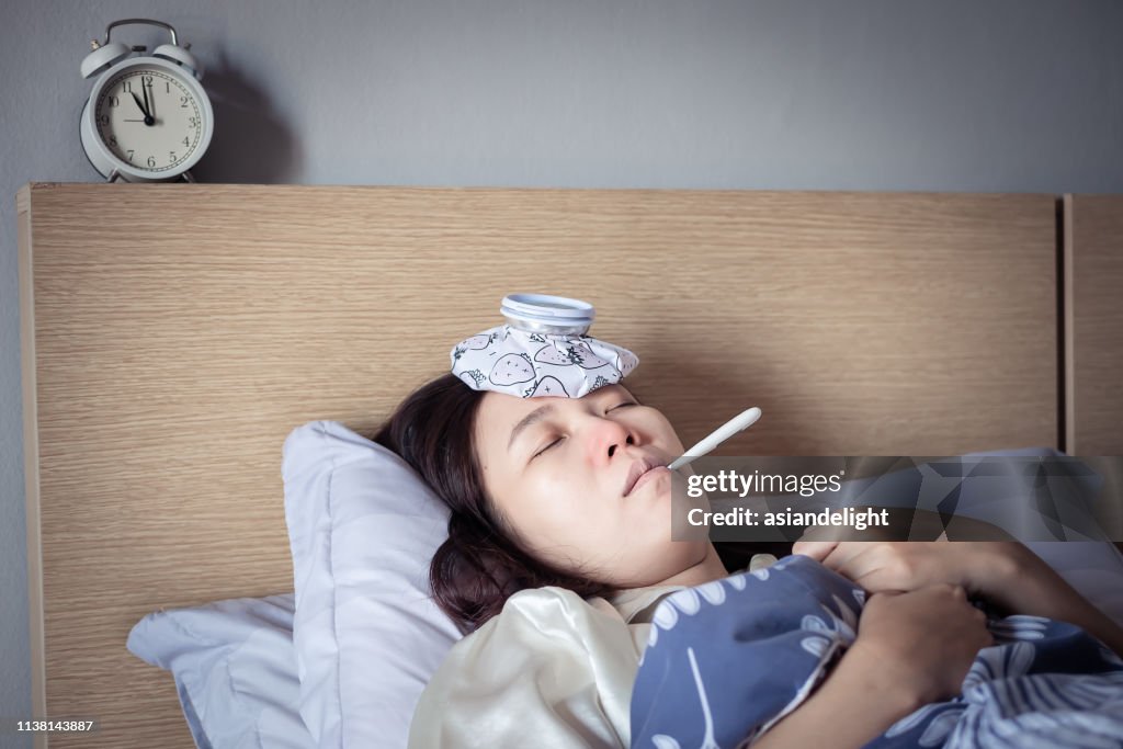 Asian woman have a cold with high fever, sleeping on cozy bed using ice bag for injuries reduce and use thermometer for measuring the temperature. sick at home