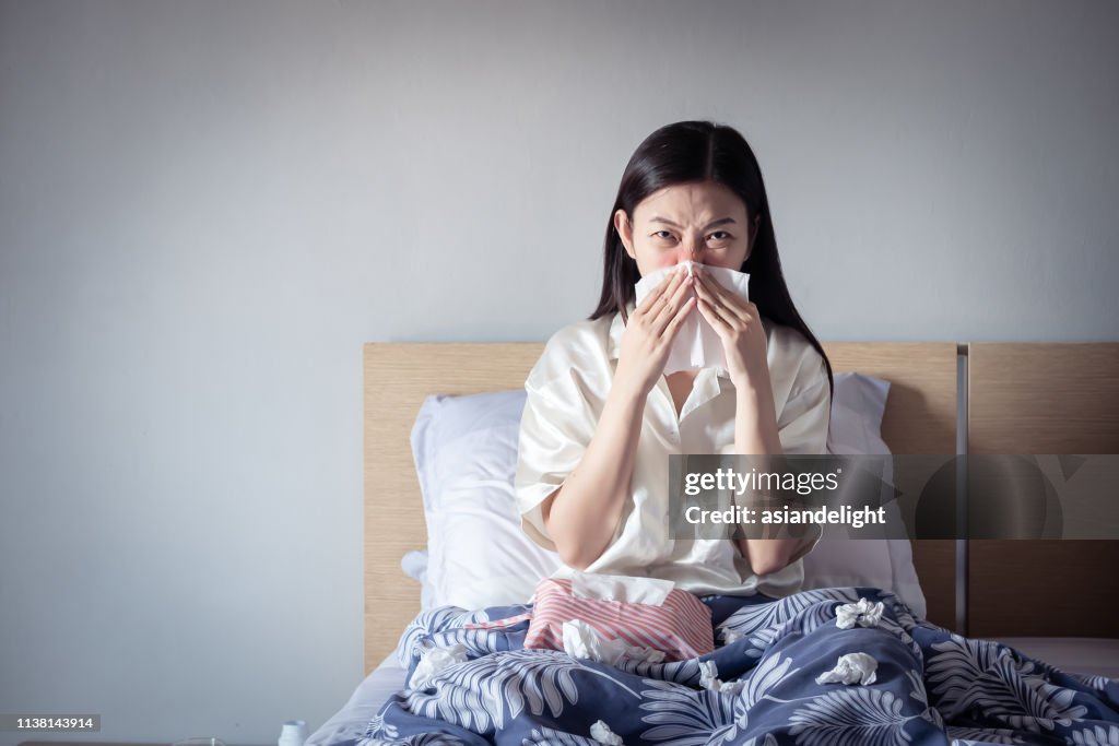 Asian woman have a cold, sitting on cozy bed using tissue for snot. sick at home