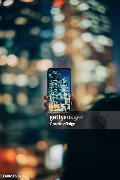 woman capturing the prosperity of city night scene with smartphone against illuminated cityscape - woman capturing city night foto e immagini stock