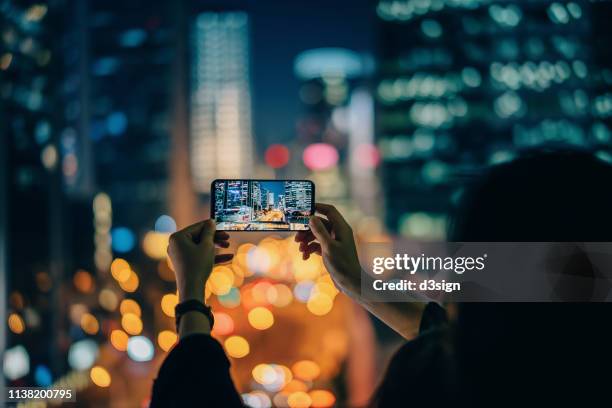 woman capturing the prosperity of city night scene with smartphone against illuminated cityscape - woman capturing city night foto e immagini stock