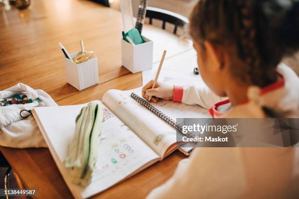 rear view of girl writing homework on table while sitting at home - compito a casa fotografías e imágenes de stock