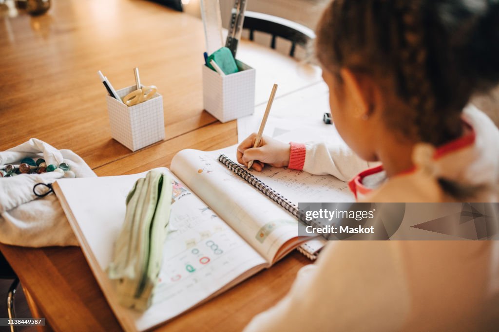 Rear view of girl writing homework on table while sitting at home