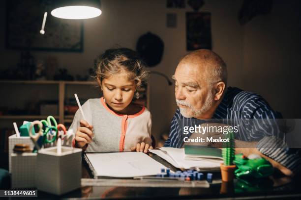 grandfather looking at granddaughter writing homework on desk in house - young girls homework stockfoto's en -beelden